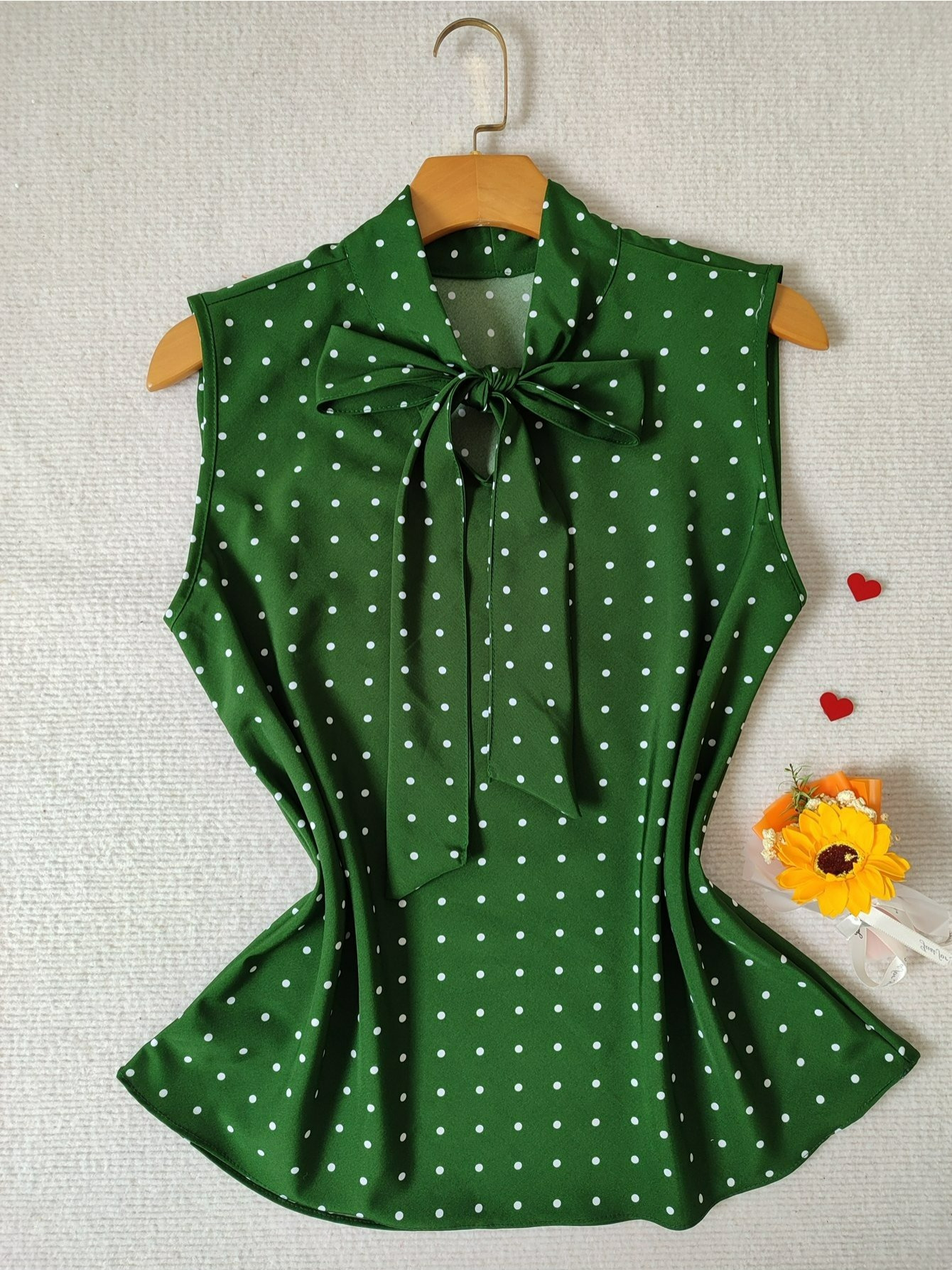 Regata plus size verde com estampa de bolinhas brancas, gola com laço e cabide de madeira, exibida sobre fundo texturizado com decoração de girassol