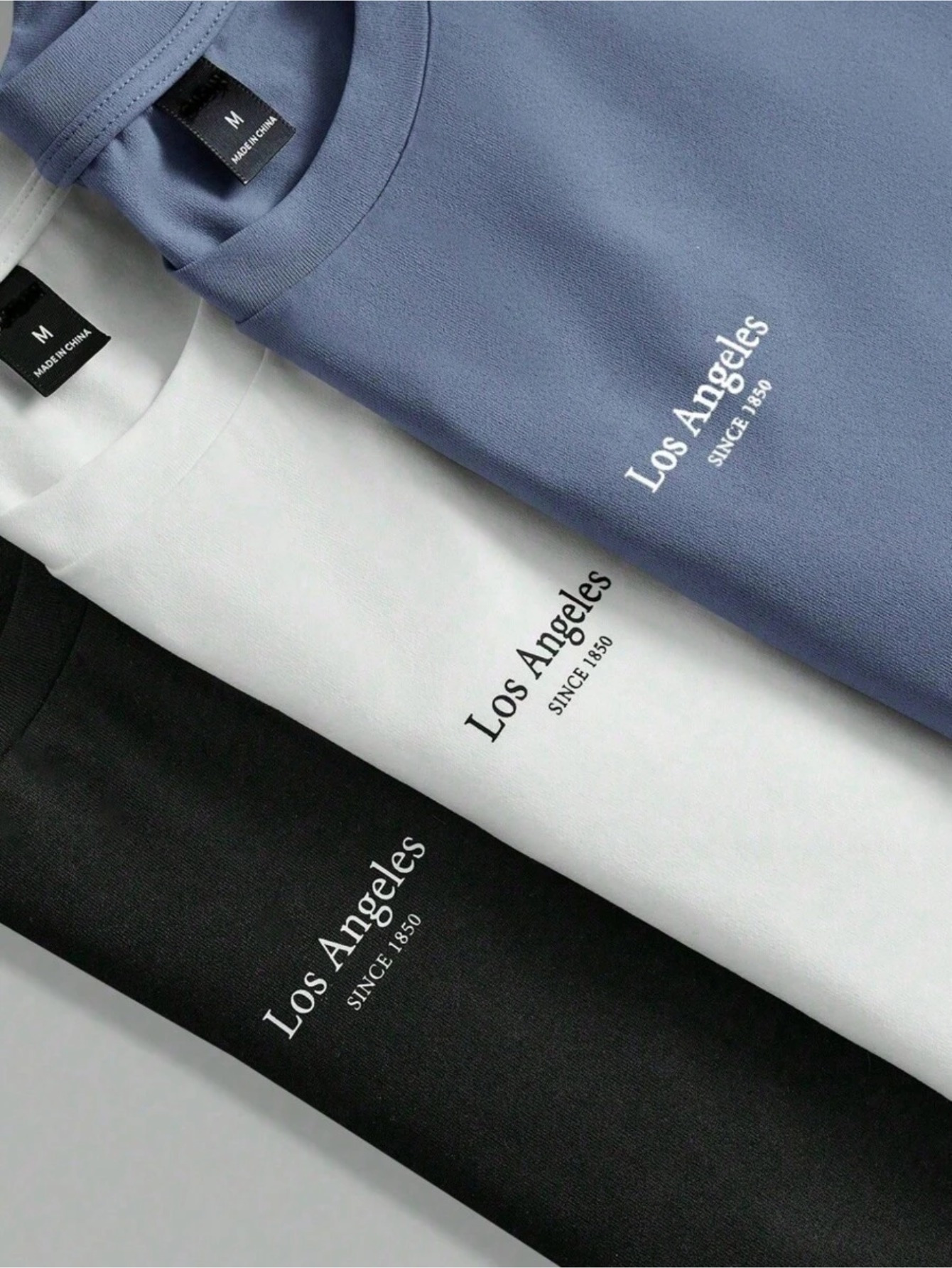 Detalhe das três camisetas sobrepostas exibindo a estampa Los Angeles SINCE 1850 e etiquetas