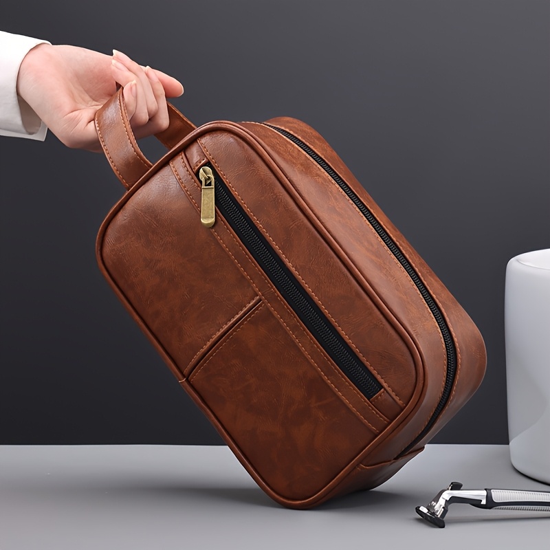 A hand holding a brown leather toiletry bag sold by 100 Spartans.