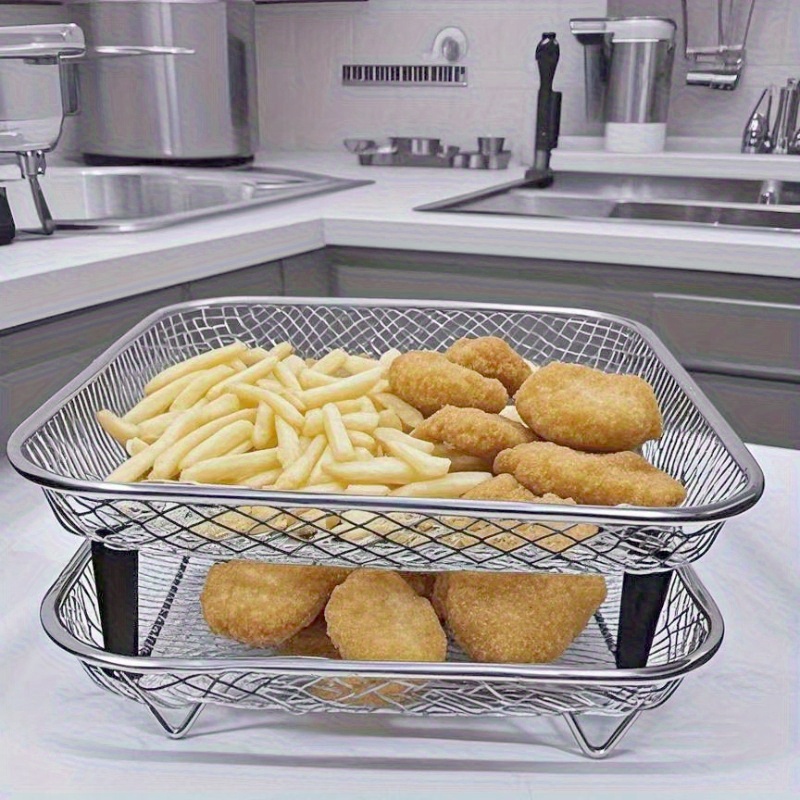 Duas bandejas aramadas de aço inoxidável empilhadas em uma cozinha. A bandeja superior contém batatas fritas e nuggets de frango, enquanto a bandeja inferior contém mais nuggets. O conjunto demonstra o uso de prateleiras duplas para otimizar o espaço e a circulação de ar durante o preparo de alimentos em fritadeiras ou fornos.